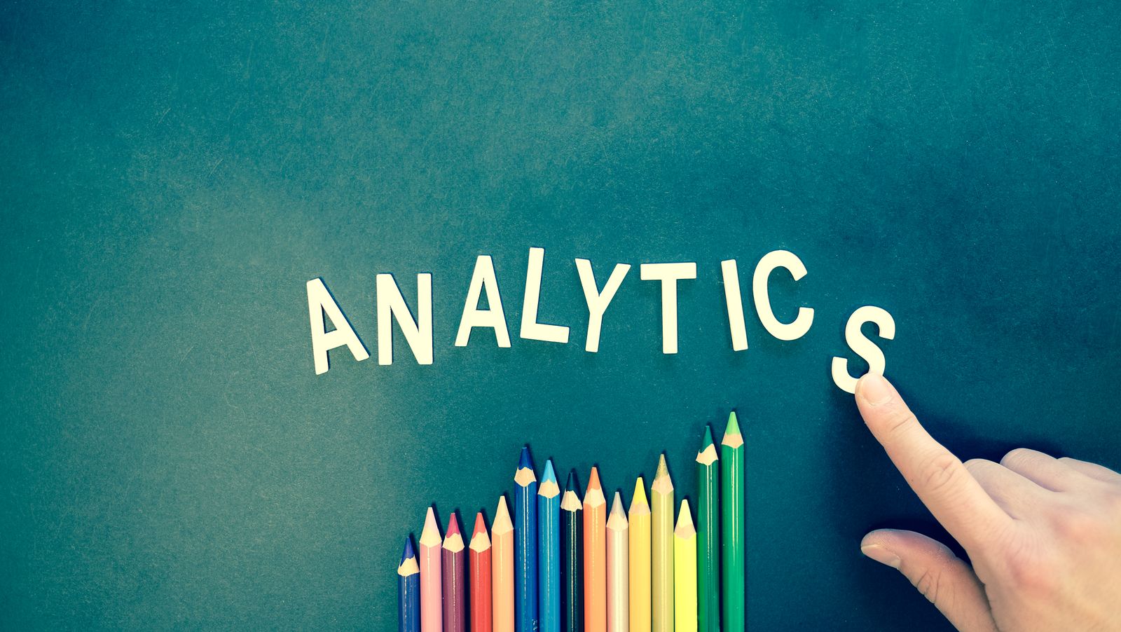 A palavra ANALYTICS em letras recortadas acima de uma fileira de lápis coloridos, com uma mão apontando.